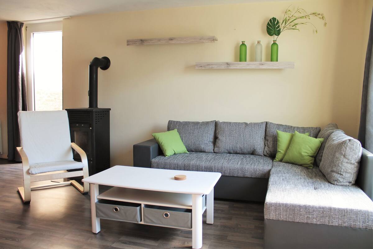 Un salon avec un fauteuil blanc, un canapé gris agrémenté de coussins verts, une table basse blanche et une cheminée.
