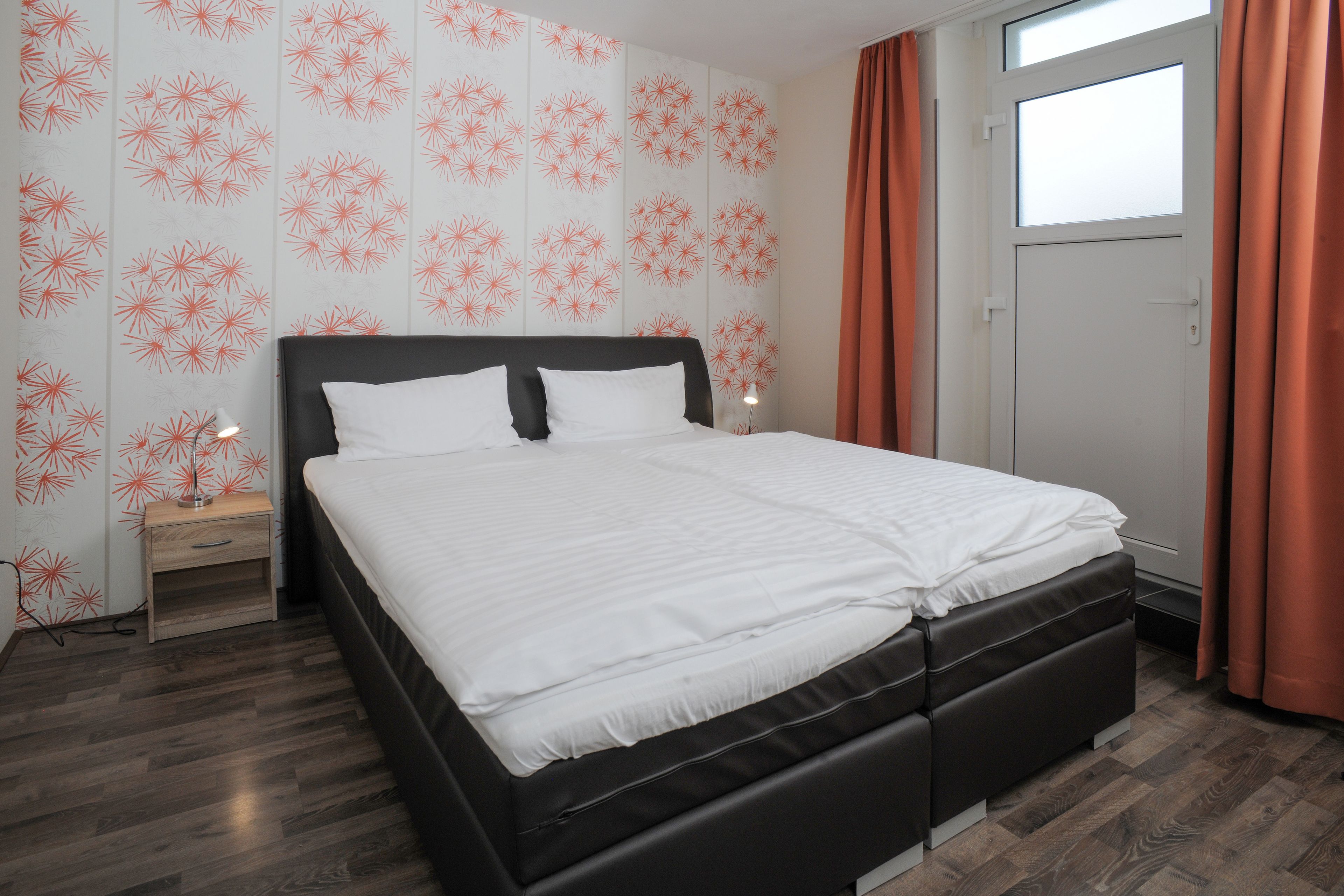 Una cama bien hecha en una habitación con papel tapiz estampado, una mesita de noche y una ventana con cortinas.