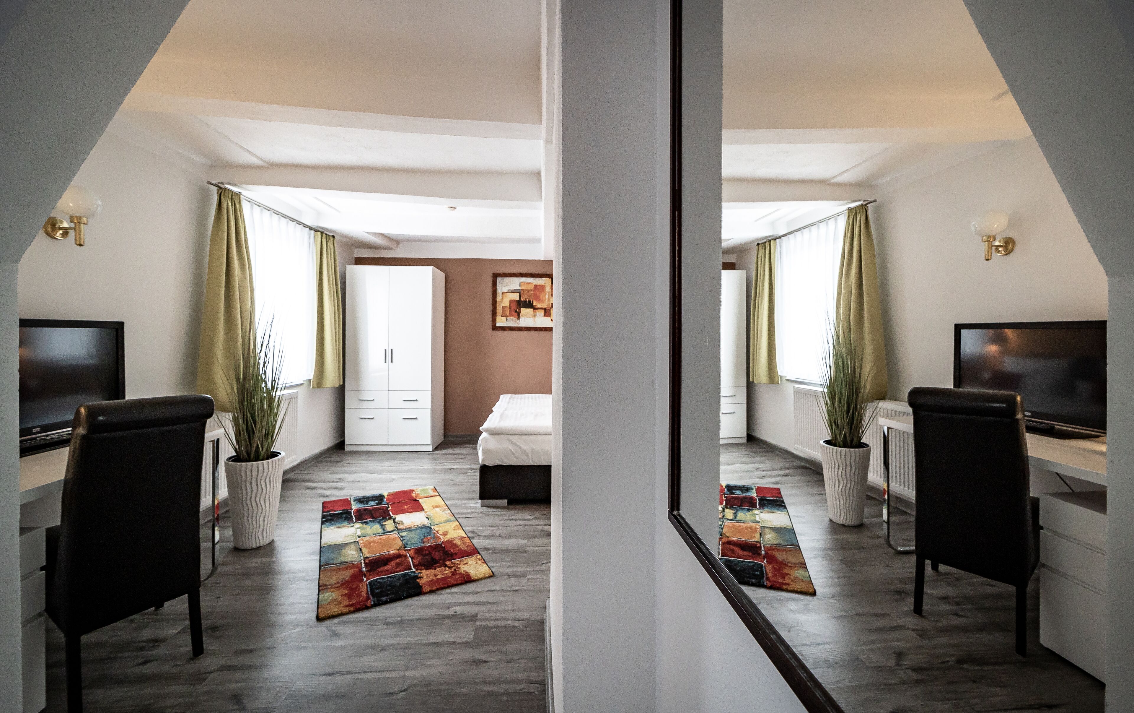 Ein Spiegelbild zeigt ein modernes Schlafzimmer mit Bett, Nachttischen, einem Fernseher und einem Fenster mit Vorhängen.