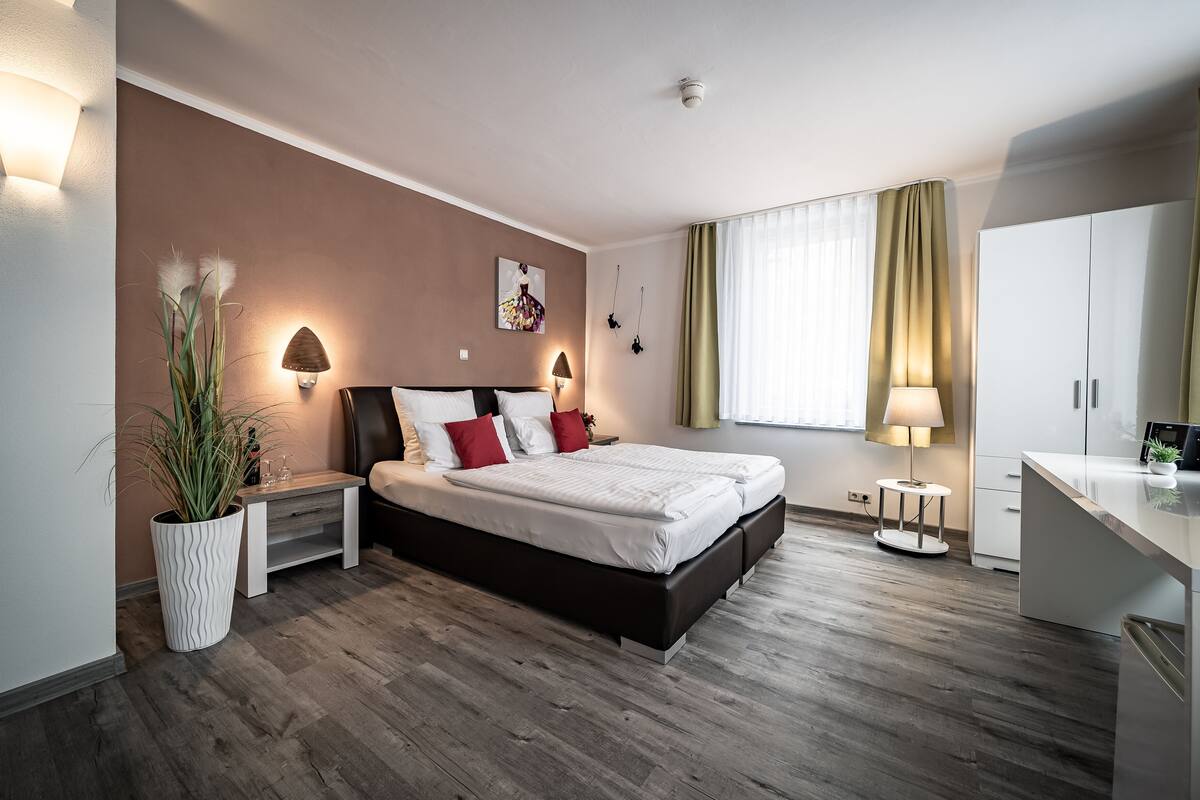 Une chambre d’hôtel moderne, équipée d’un lit, de tables de chevet, d’une plante en pot, d’un bureau et d’une fenêtre avec des rideaux.