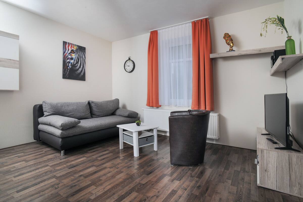 Un salon moderne avec un canapé gris, une table basse blanche, une poubelle noire, un téléviseur posé sur un meuble en bois et une fenêtre ornée de rideaux orange.