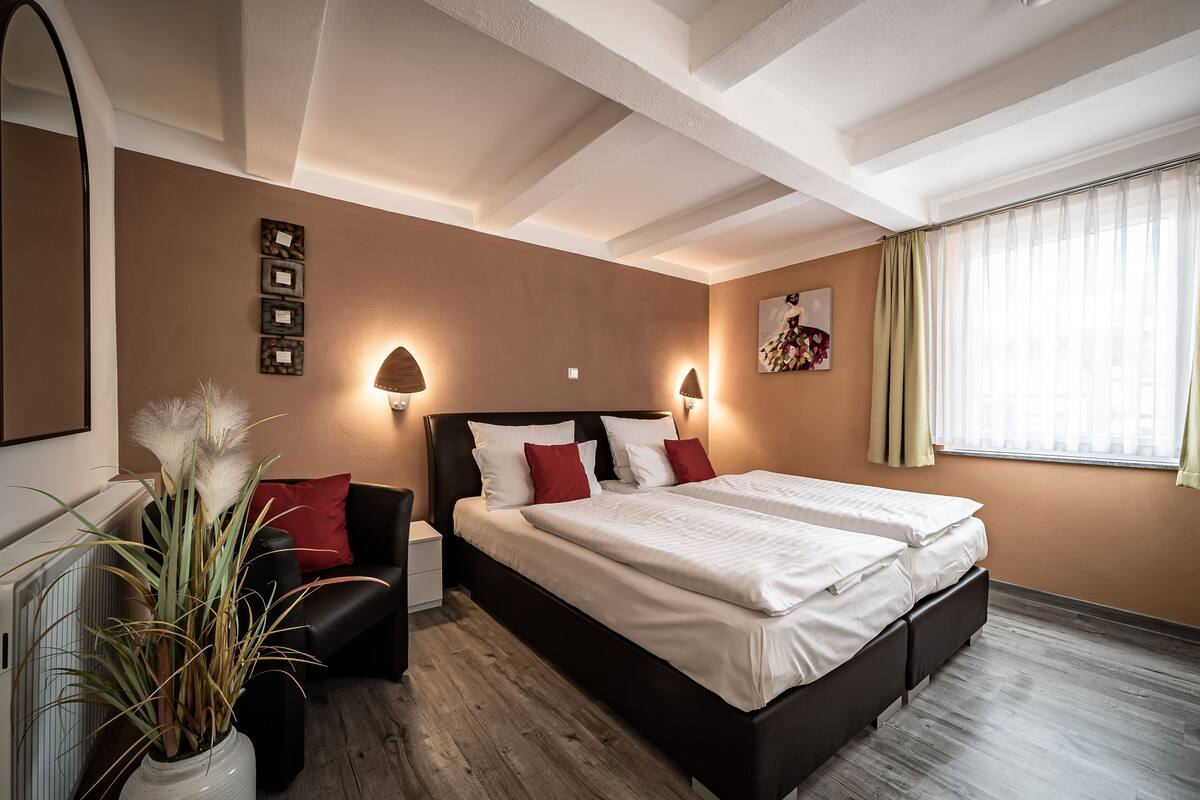 Une chambre d’hôtel avec un grand lit, une table de chevet, une lampe, une plante en pot et une fenêtre avec des rideaux.