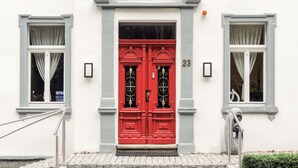 Ein Gebäude mit roter Tür, Hausnummer 23, und zwei mit Vorhängen versehene Fenster.