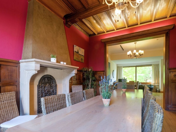 Une salle à manger élégante, avec une longue table, des chaises en osier, une cheminée et un lustre.