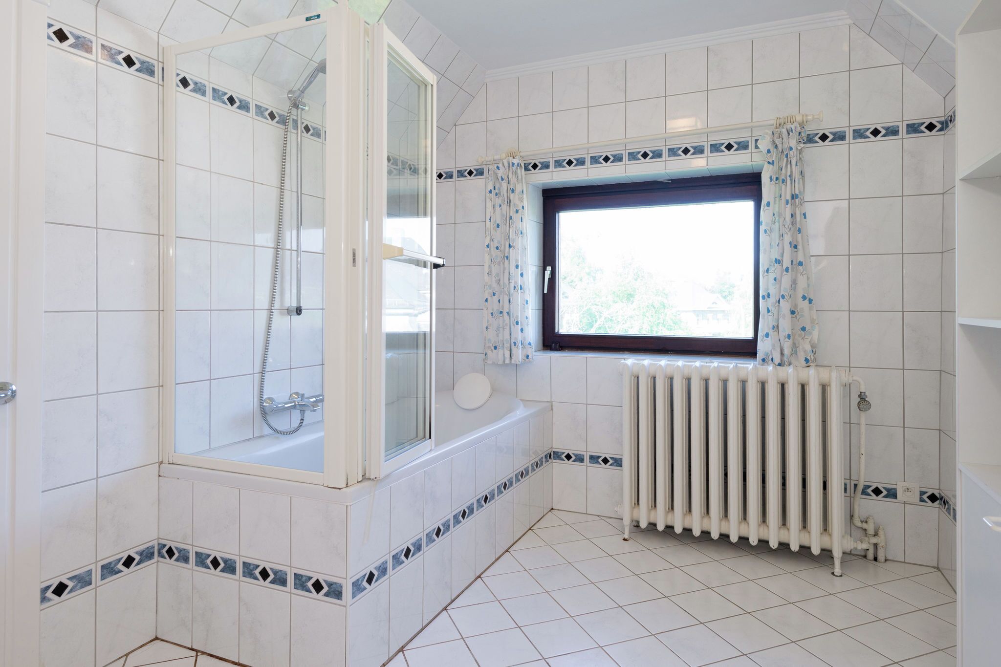 Un baño con paredes de azulejos blancos, cabina de ducha, bañera, ventana con cortina y radiador.
