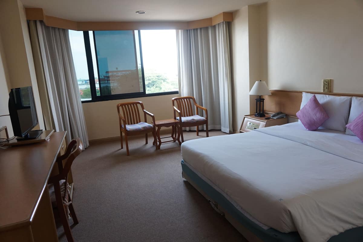 Habitación de hotel con una cama grande, un escritorio con silla, una mesita de noche con lámpara y una ventana con cortinas.