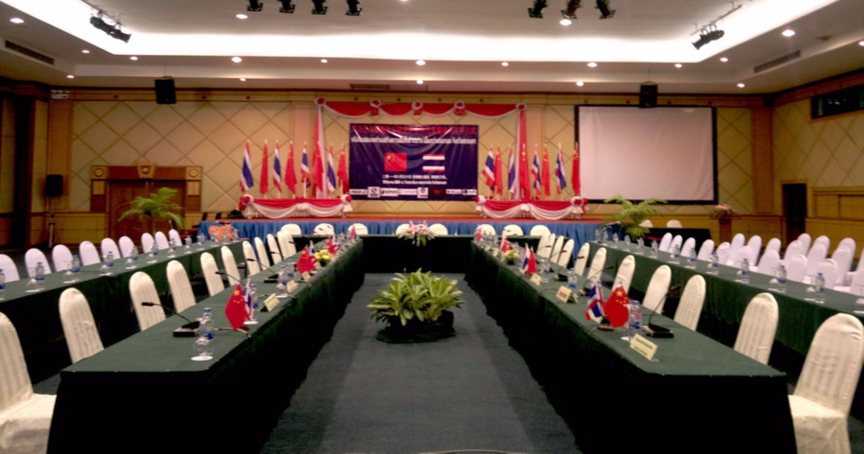 Une salle de conférence avec une longue table, des chaises blanches et une estrade décorée de drapeaux.