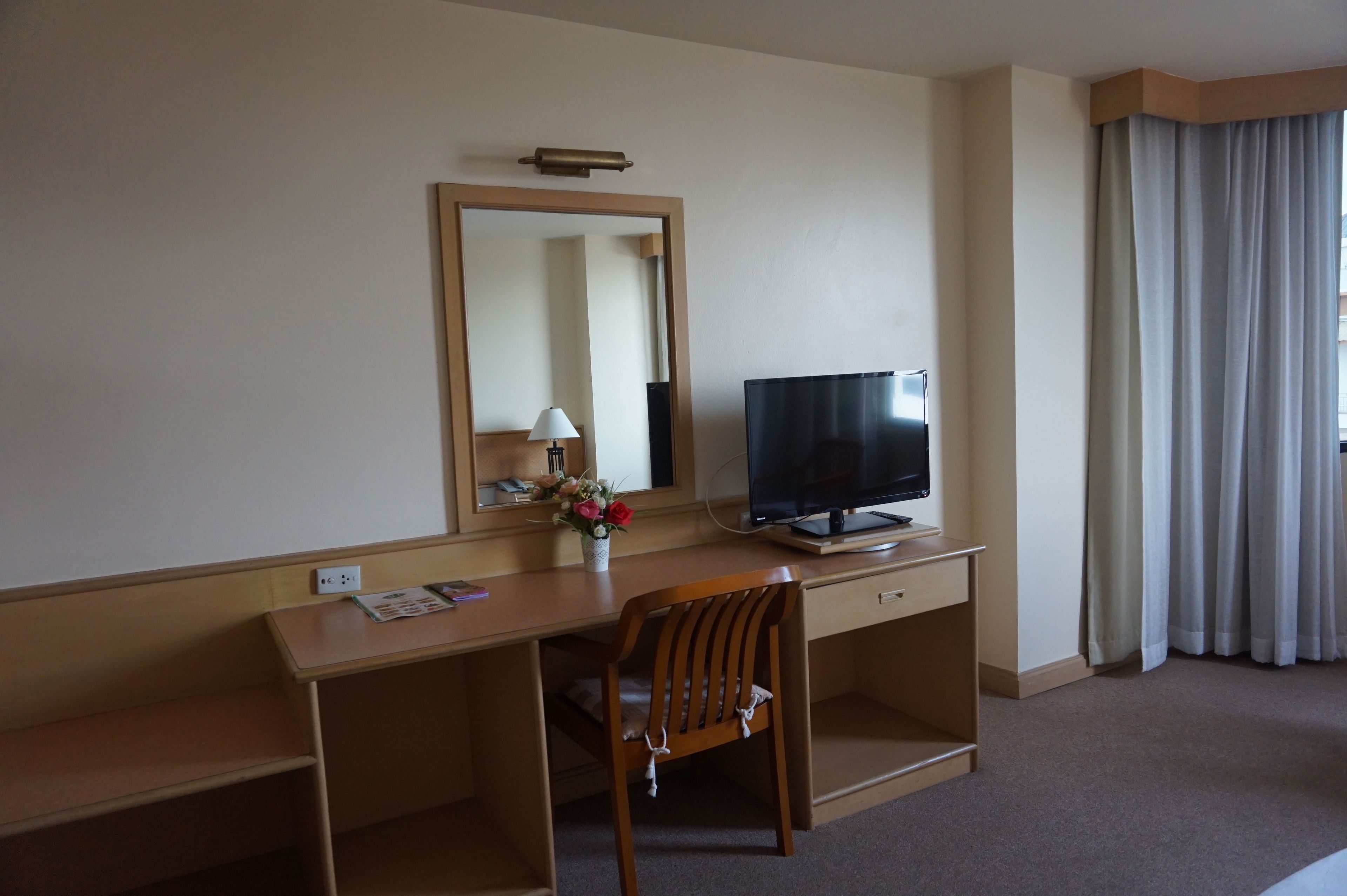 Une chambre d’hôtel avec un bureau en bois, une télévision, un miroir, une chaise et une fenêtre avec des rideaux.