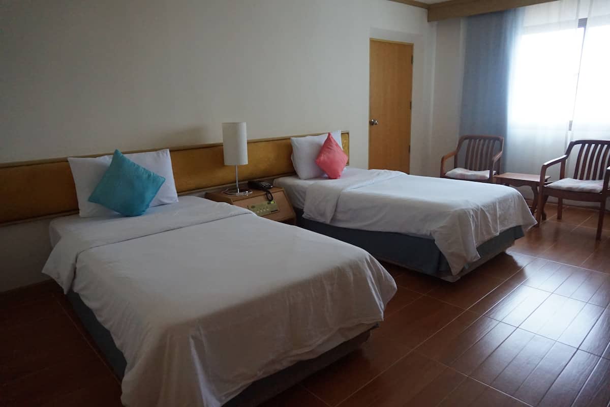 Habitación de hotel con dos camas, una mesita de noche con lámpara, una silla y una ventana con cortinas.