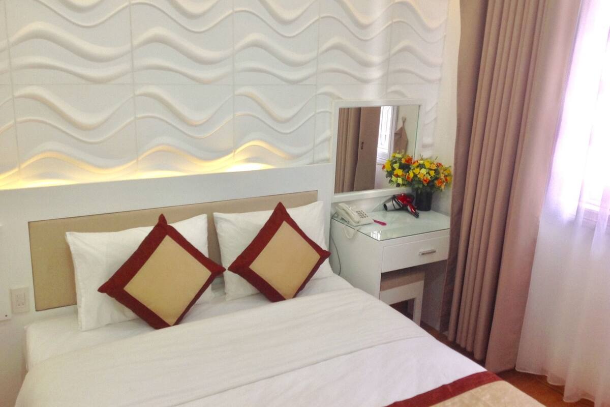 Una habitación de hotel con una cama, una mesita de noche con teléfono, un espejo y un jarrón con flores.