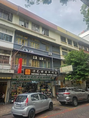 A multi-story building with a variety of shops on the ground floor, including a supermarket. There are parked cars on the street in front.