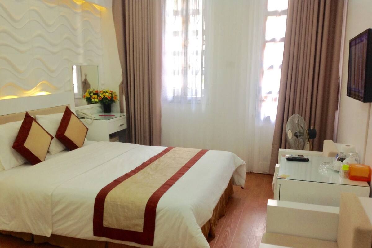 A hotel room with a large bed, a bedside table with a lamp, a small table with a fan, and a TV on the wall.