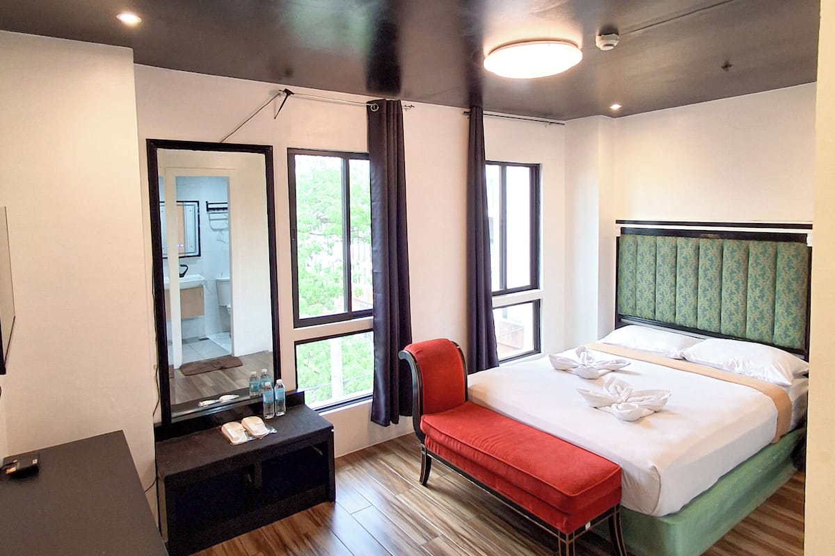A hotel room with a bed, a desk, a chair, a mirror, and large windows.