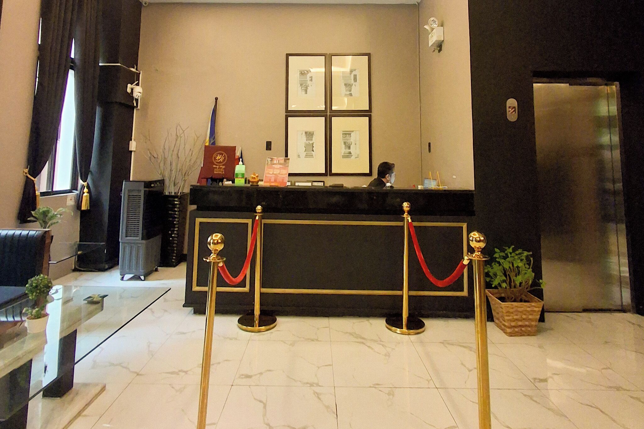 Zona de recepción de un hotel con suelo de mármol, mostrador de recepción, cuadros enmarcados en la pared y un refrigerador.