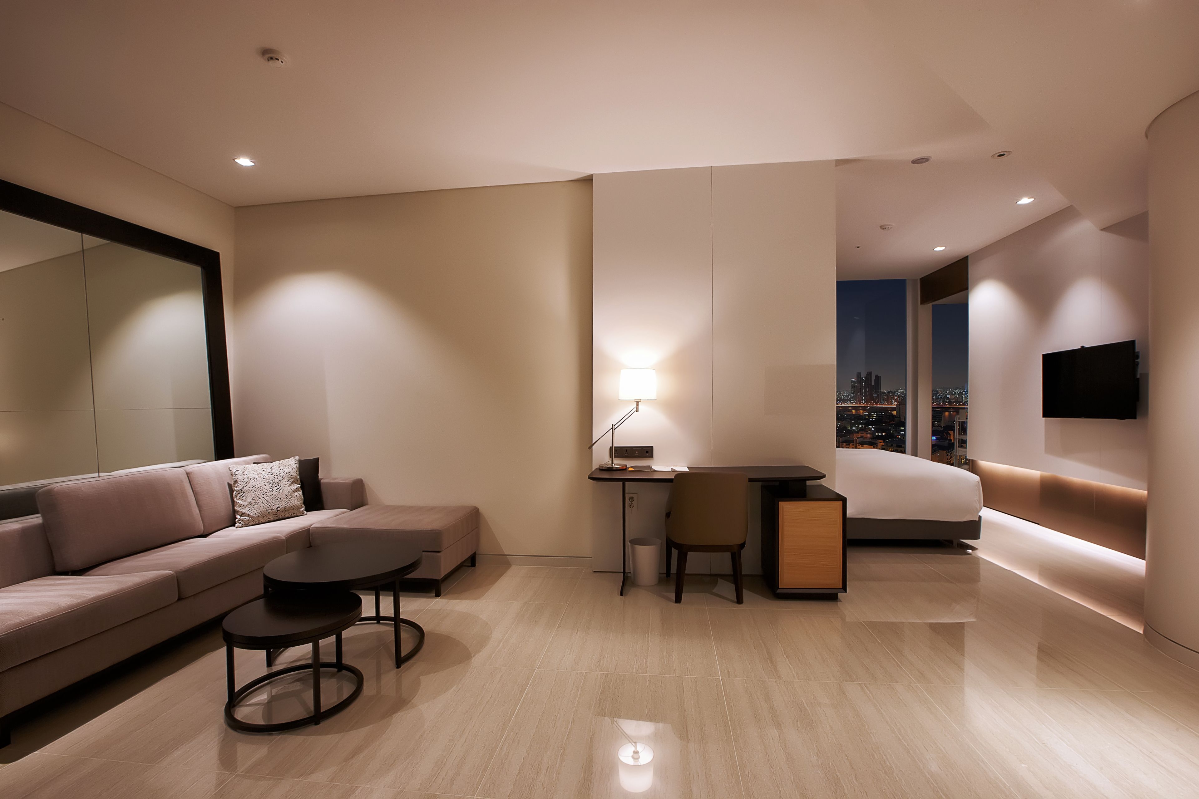 Un salon moderne avec un canapé, une petite table et un bureau avec une chaise. Un téléviseur est fixé au mur et une fenêtre offre une vue sur la ville.