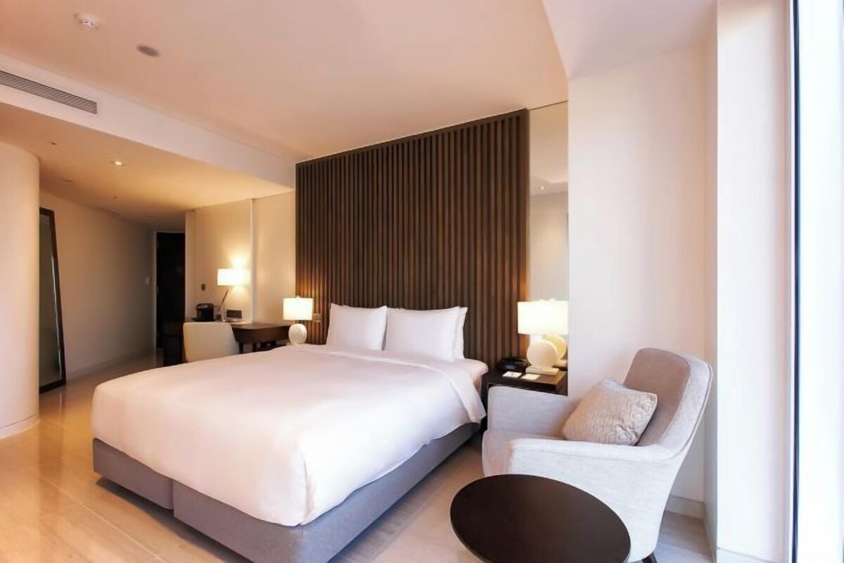 A modern hotel room with a large bed, bedside tables with lamps, a seating area with a chair, and a large window.