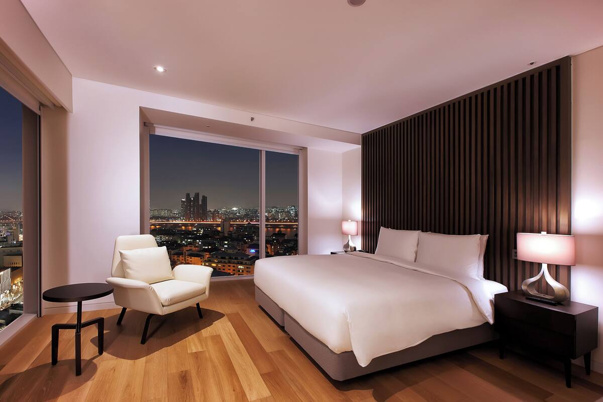 Habitación de hotel moderna con una cama grande, una silla, una mesita y una mesita de noche con una lámpara.