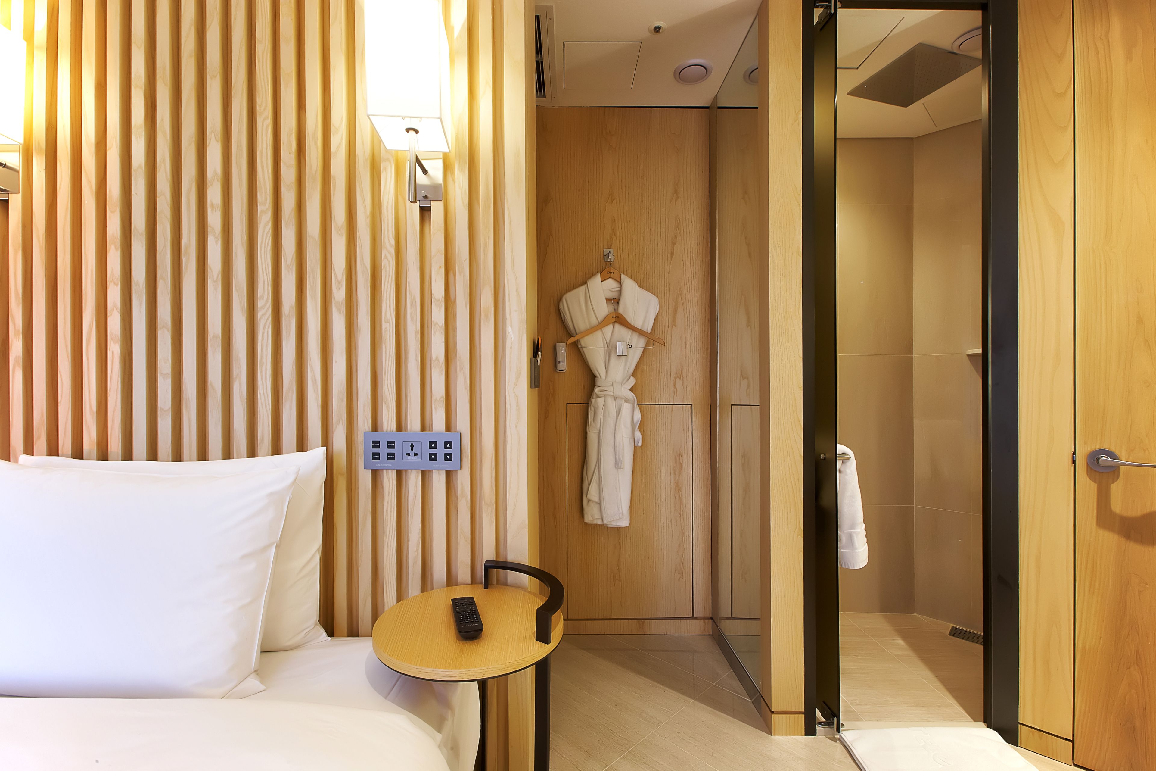 Habitación de hotel con cama, mesita de noche con control remoto, toma de corriente en la pared y armario de madera.