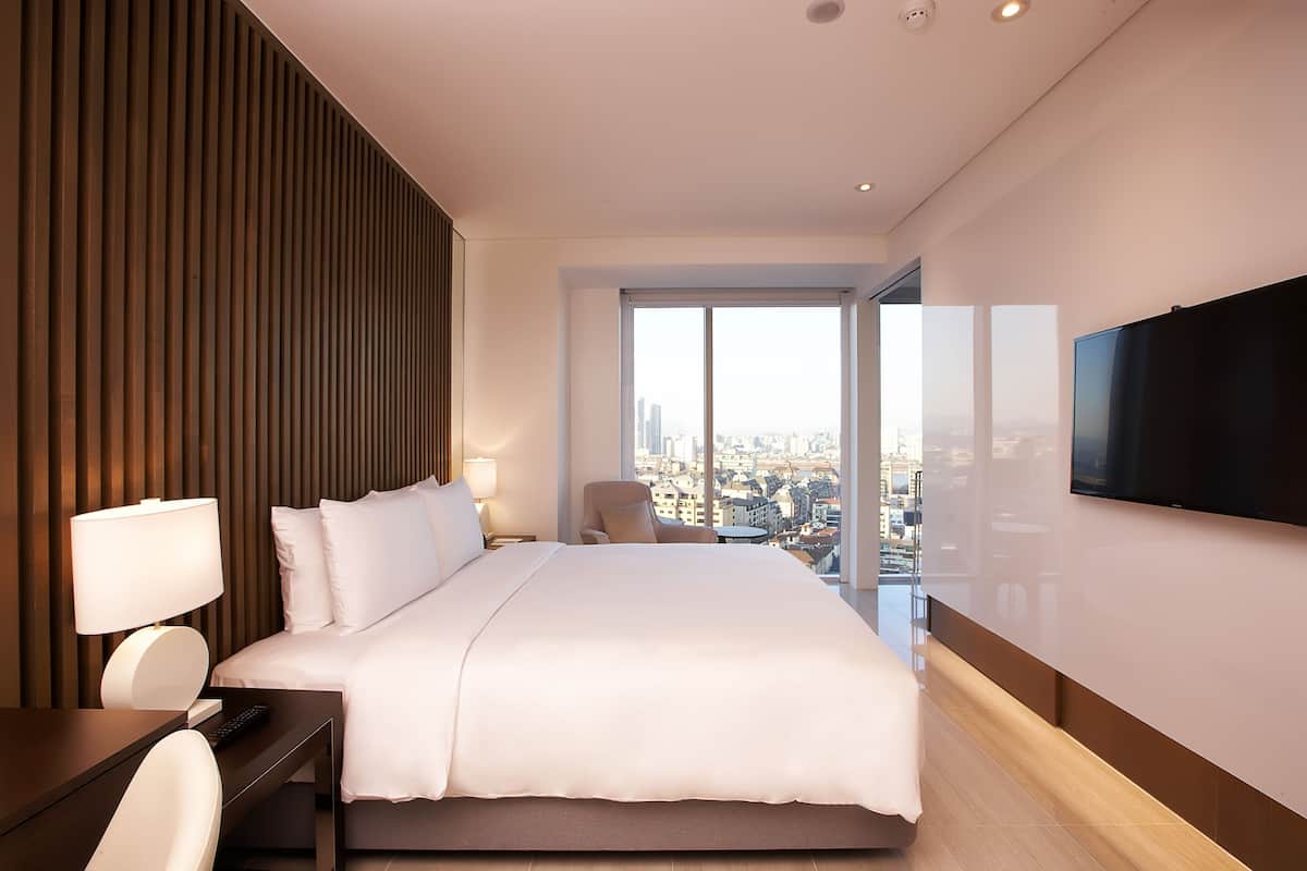 Una habitación de hotel moderna con una cama grande, un escritorio con una silla, un televisor montado en la pared y vistas a la ciudad desde la ventana.