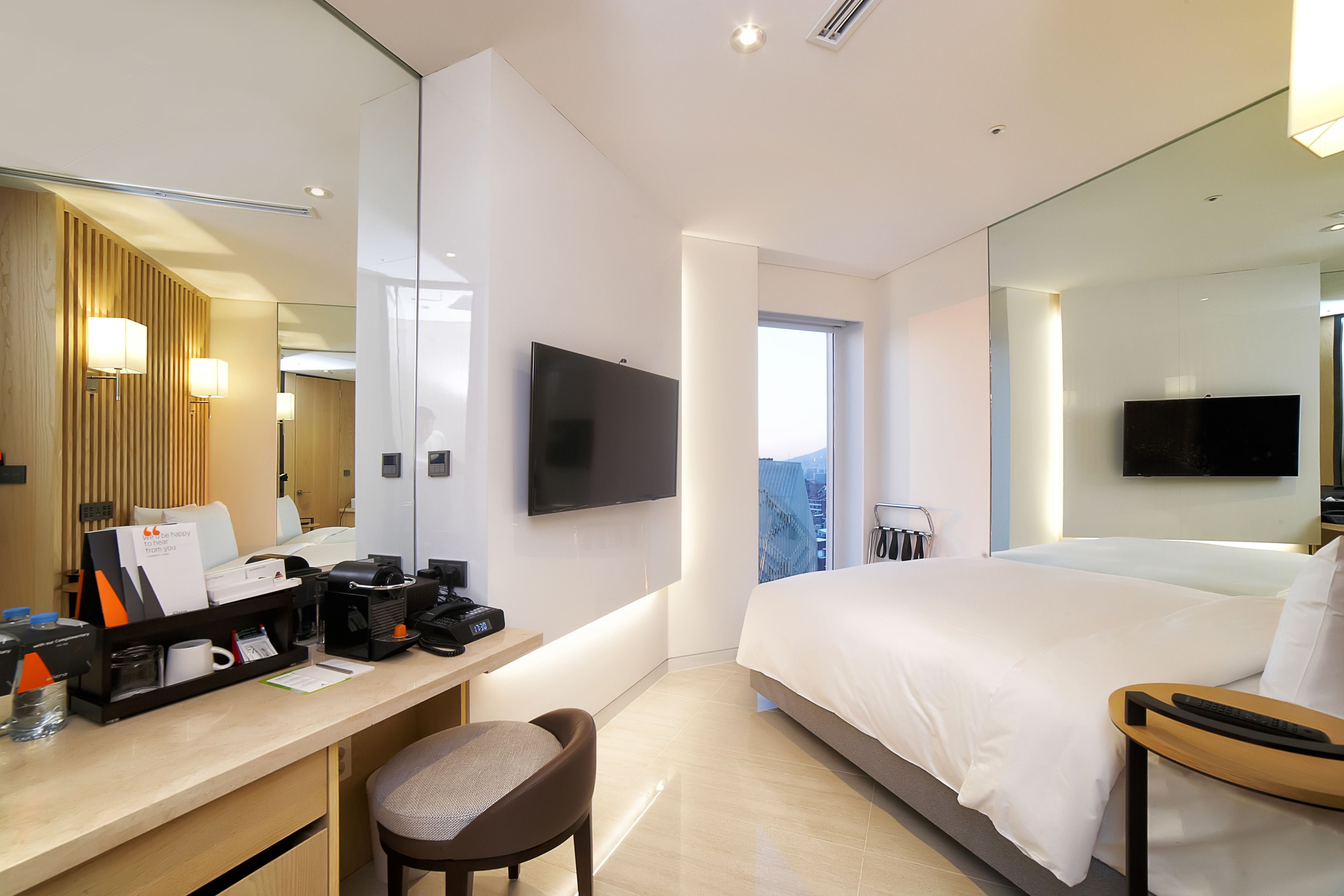 A modern hotel room with a large bed, a desk with a chair, a flat-screen TV mounted on the wall, and a large mirror reflecting the room.