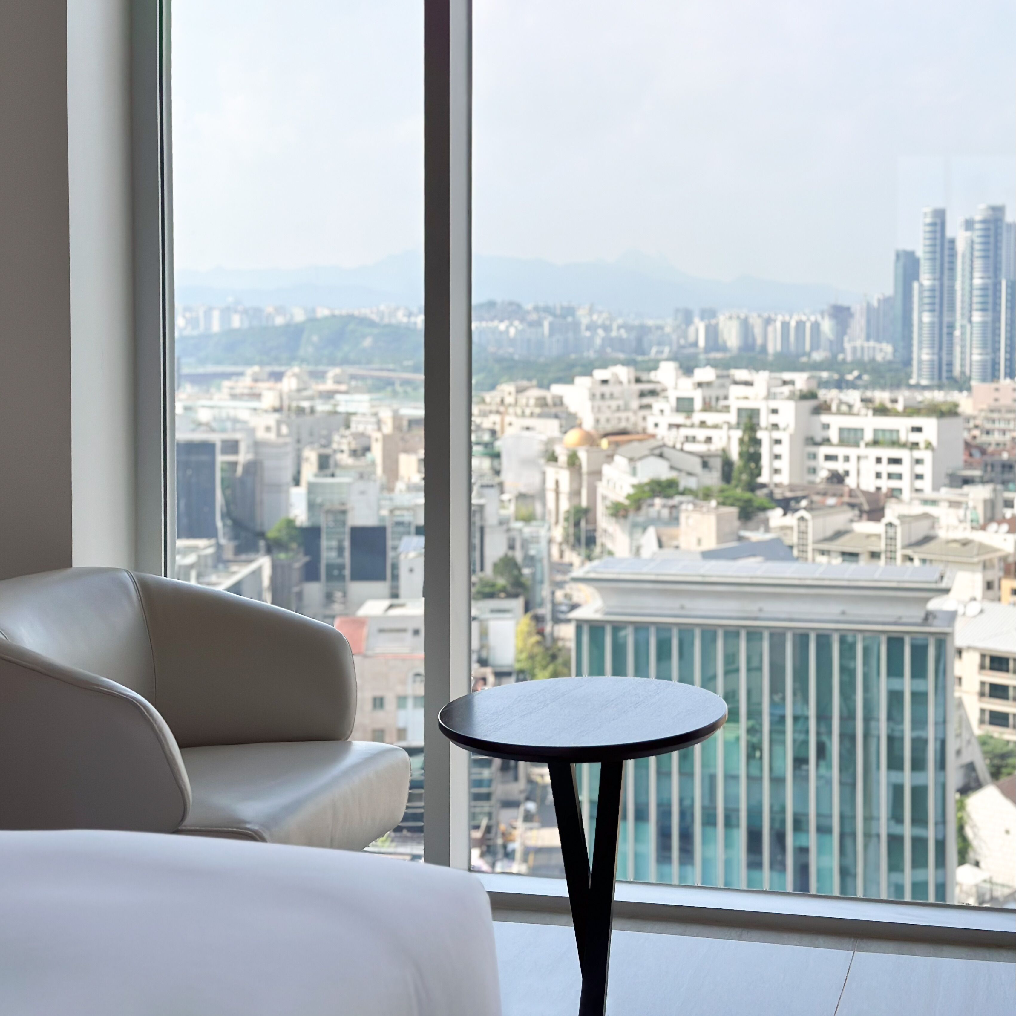 Habitación de hotel moderna con un gran ventanal que ofrece vistas a la ciudad, un sofá blanco y una pequeña mesa redonda.