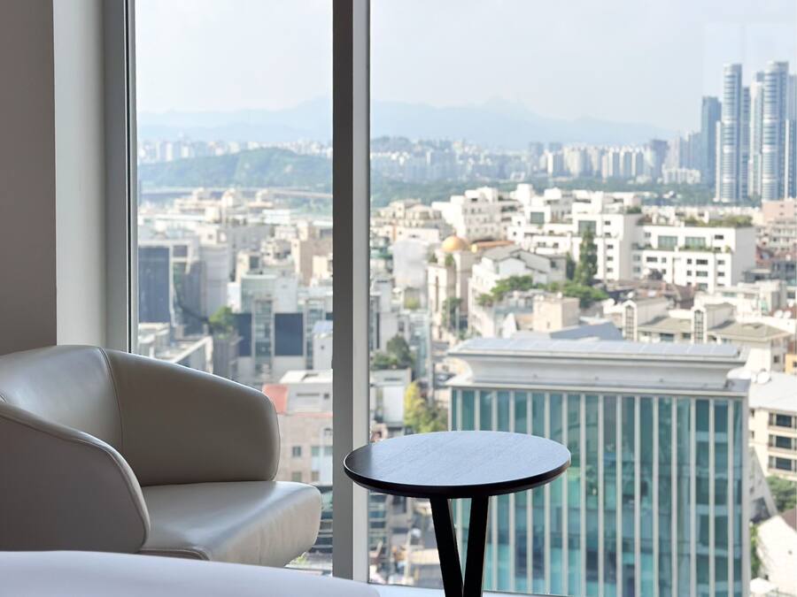 Habitación de hotel moderna con un gran ventanal que ofrece vistas a la ciudad, un sofá blanco y una pequeña mesa redonda.