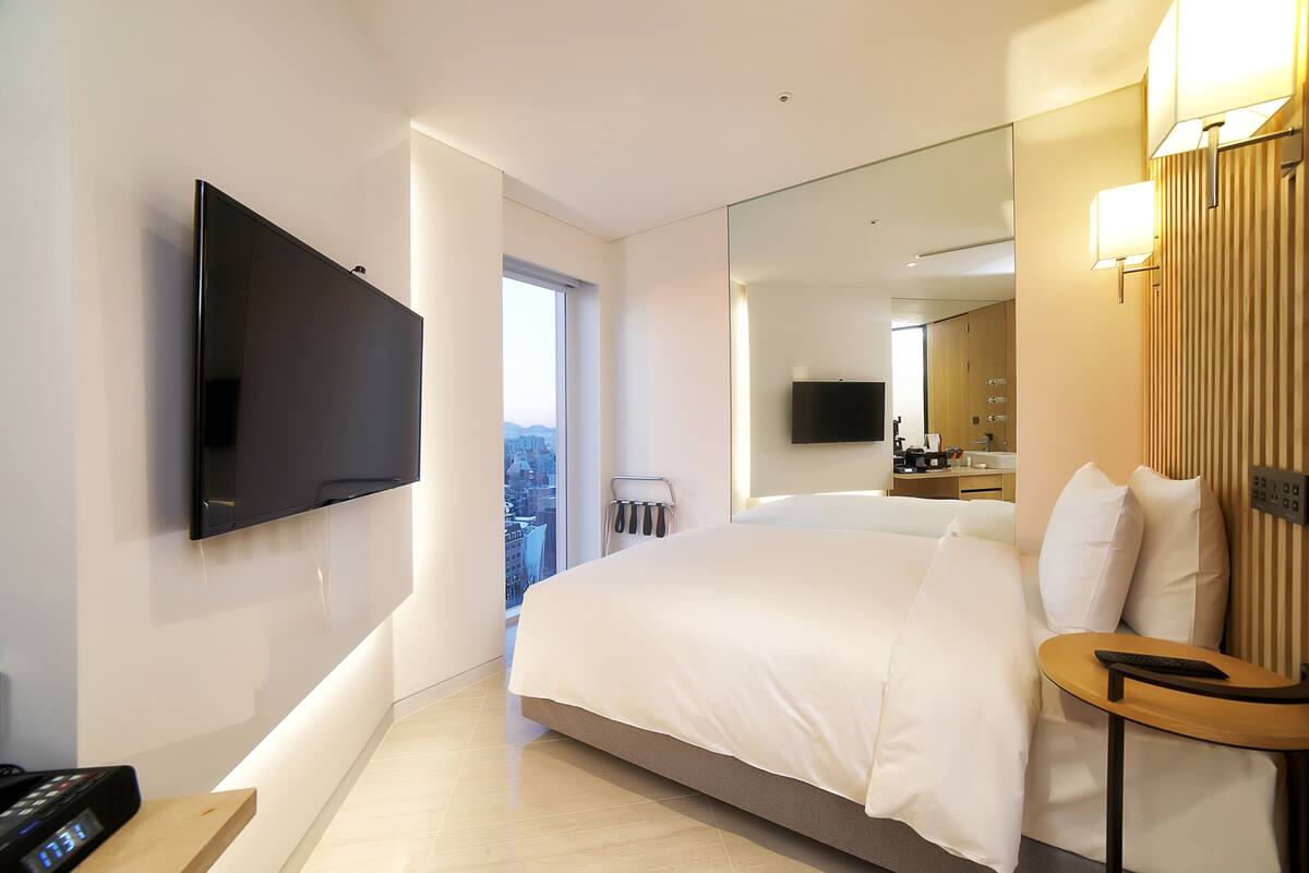 Una habitación de hotel moderna con una cama grande, un televisor de pantalla plana montado en la pared, una mesita de noche con un control remoto y un amplio ventanal con vistas a la ciudad.