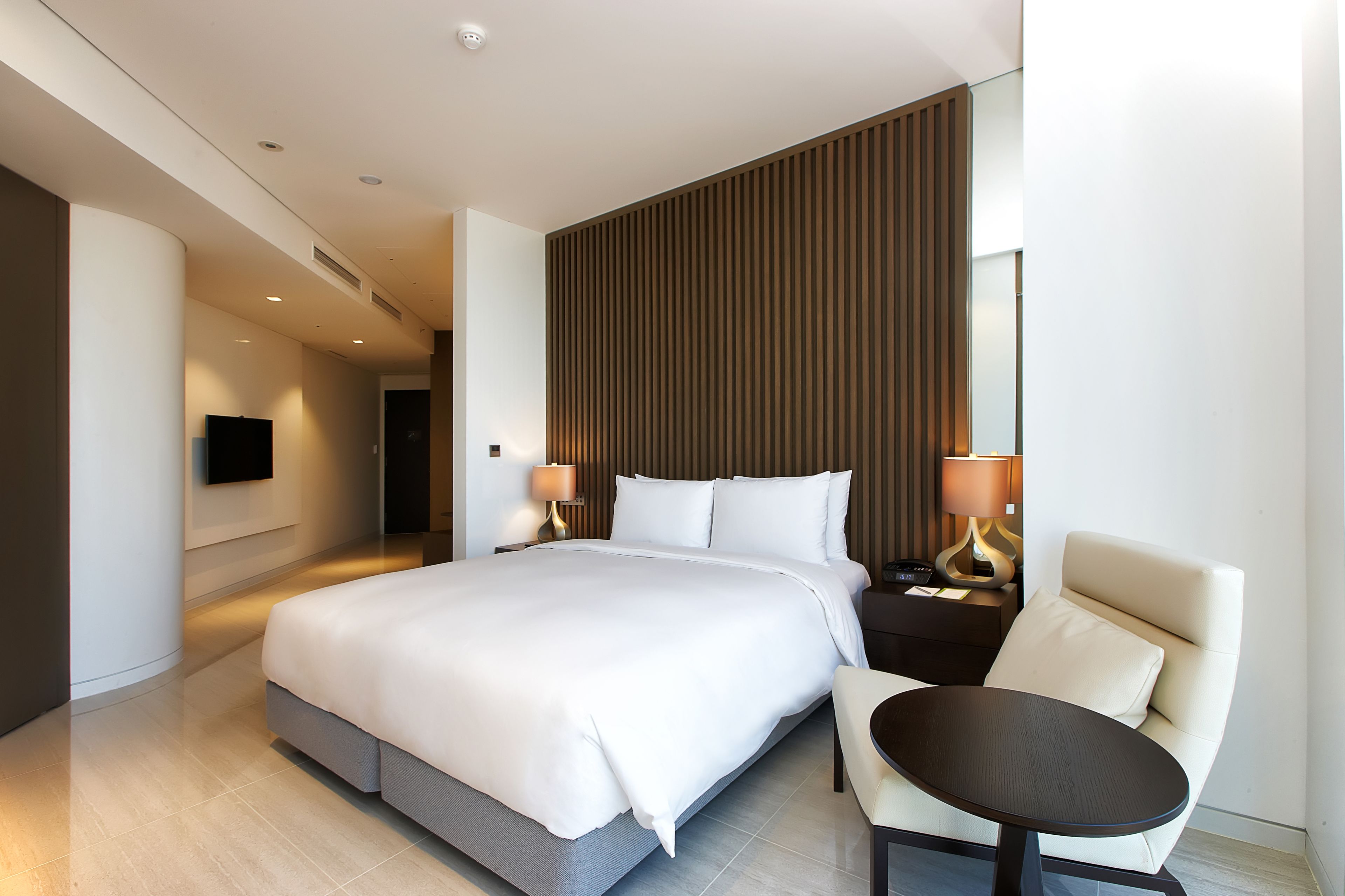Quarto de hotel moderno com uma cama grande, mesa de cabeceira com luminárias, uma poltrona e uma TV de tela plana na parede.