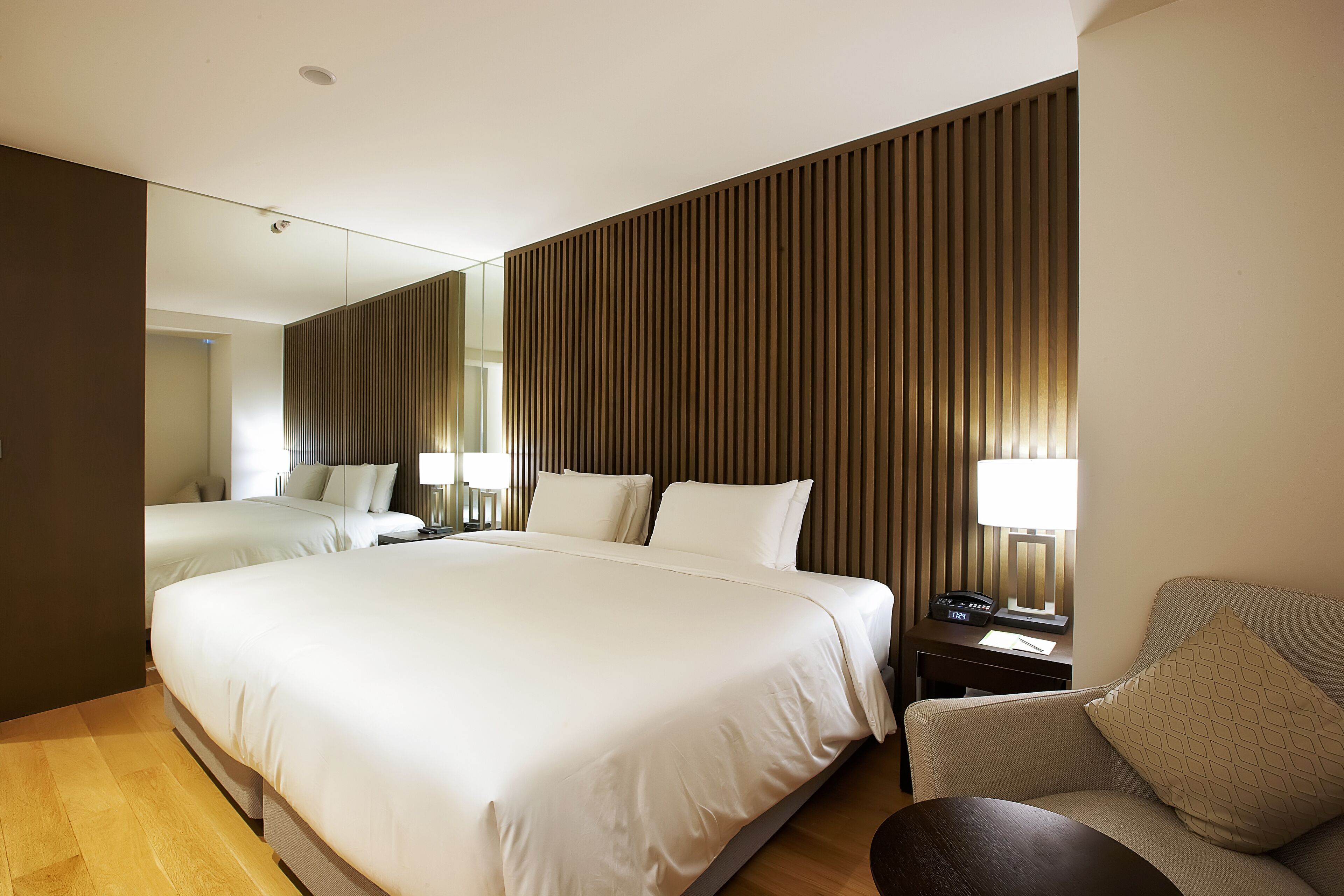 A modern hotel room with a large bed, a sofa, a desk with a lamp, and a large mirror.