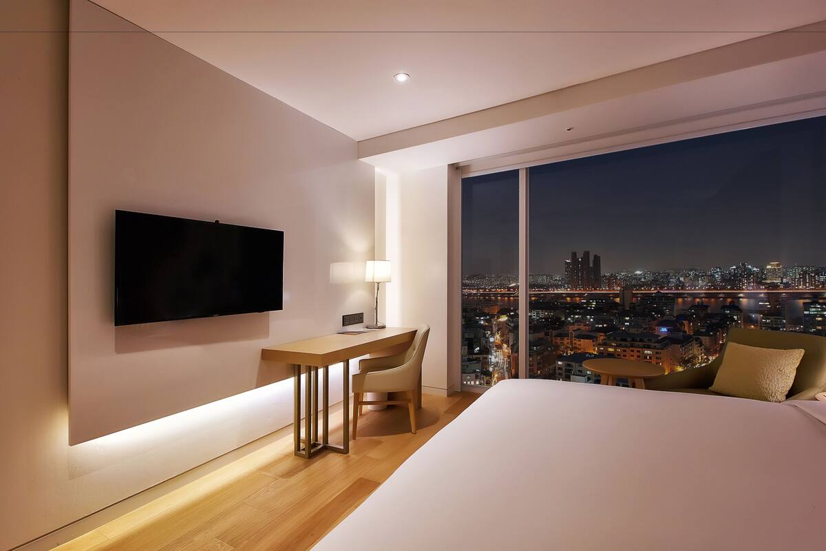 Una habitación de hotel moderna con una cama grande, un escritorio con una silla, un televisor montado en la pared y vistas a la ciudad a través de un ventanal amplio.