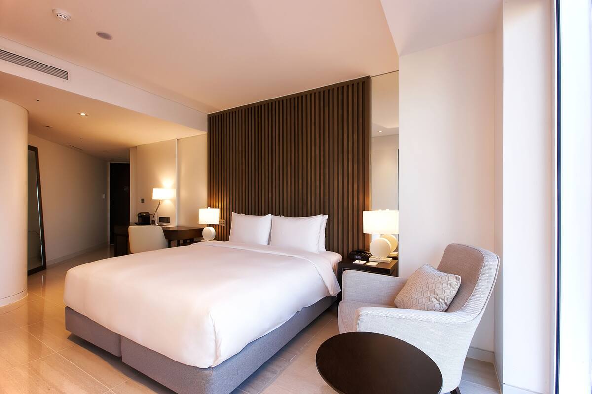 Una habitación de hotel moderna con una cama grande, una zona de estar con una silla y una mesita, y una mesita de noche con lámparas.