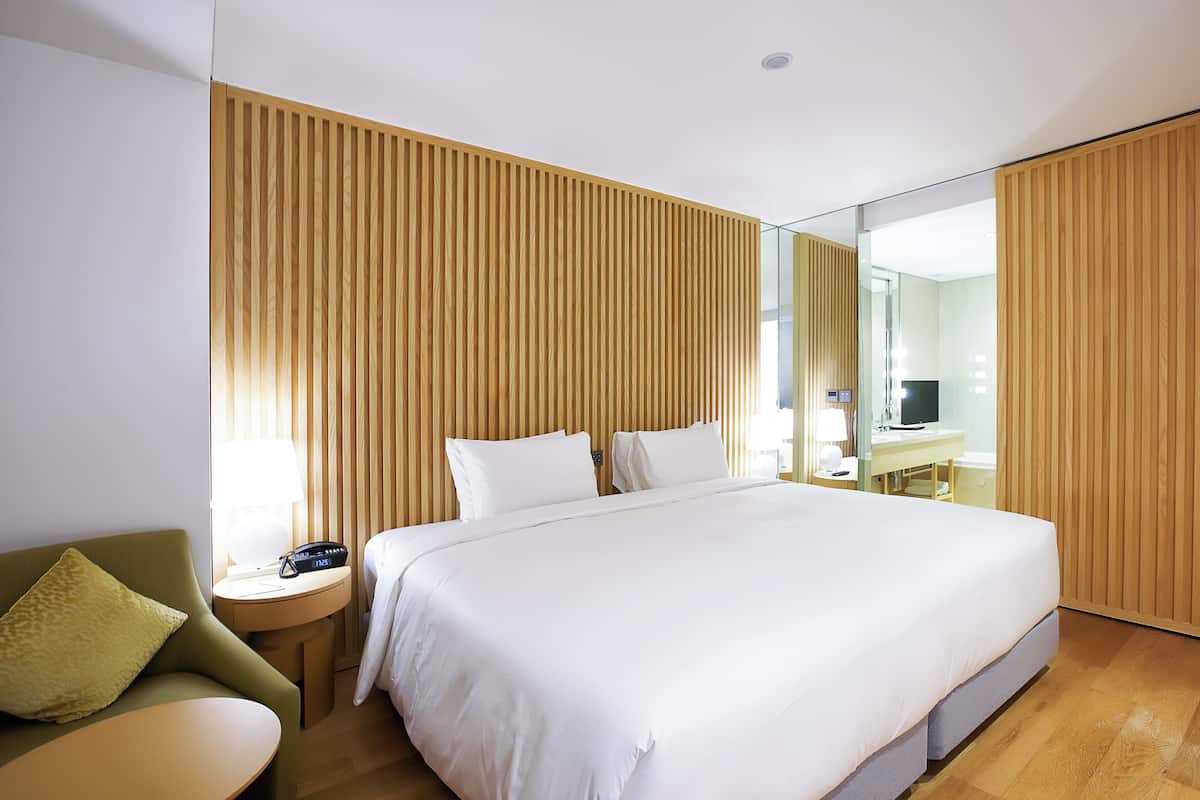 Habitación de hotel con una cama grande, una mesita de noche con lámpara, un sillón verde y un panel de pared de madera.