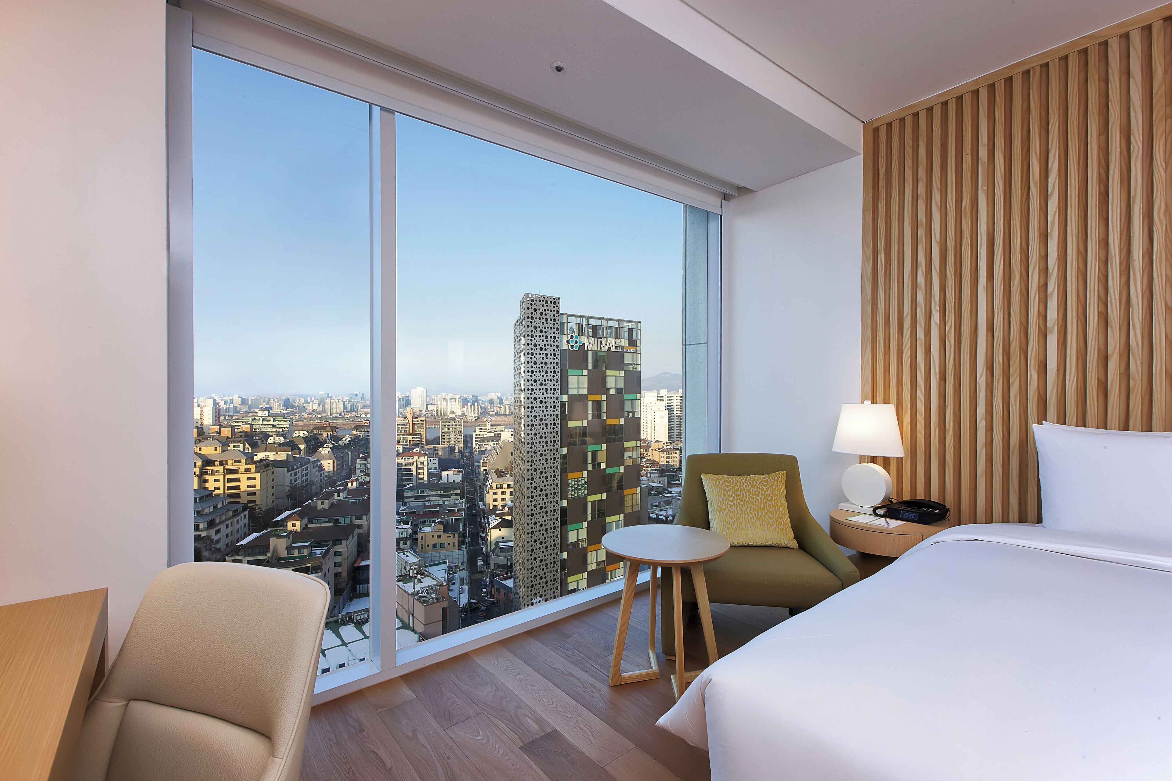 A hotel room with a large window offering a city view, a bed, a chair, a small table, and a nightstand with a lamp.