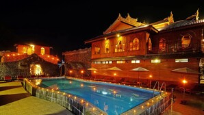 Una piscina con borde de piedra, rodeada de sillas de descanso y sombrillas, y un edificio grande con arquitectura tradicional al fondo.