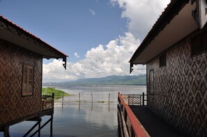 A wooden structure with a patterned exterior, leading to a body of water with mountains in the background. - Ann Heritage Lodge (Nyaungshwe)