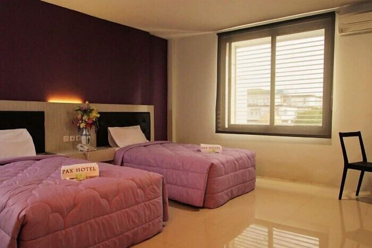 A hotel room with two beds, a window with blinds, a chair, and a vase with flowers.