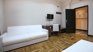 A hotel room with a white sofa, a desk with a chair, a flat-screen TV, and a door leading to a hallway with a world map. - Guesthouse Vittoria Rooms (Genoa)