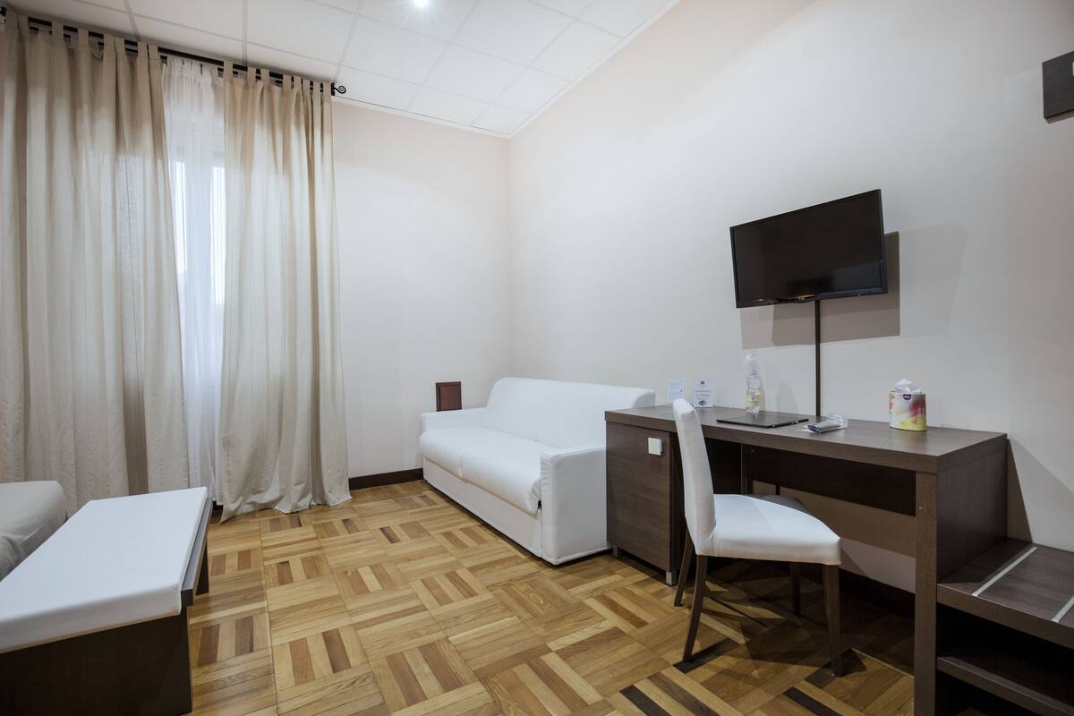 A hotel room with a wooden parquet floor, a white sofa, a desk with a chair, a flat-screen TV mounted on the wall, and a window with white curtains.