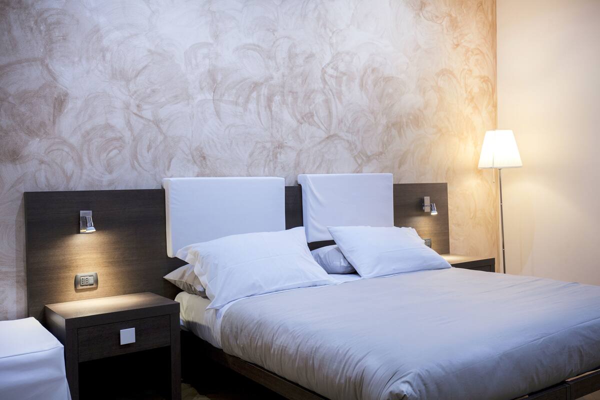 A neatly made bed with white linens and pillows, a bedside table with a lamp, and a wall with a textured pattern.