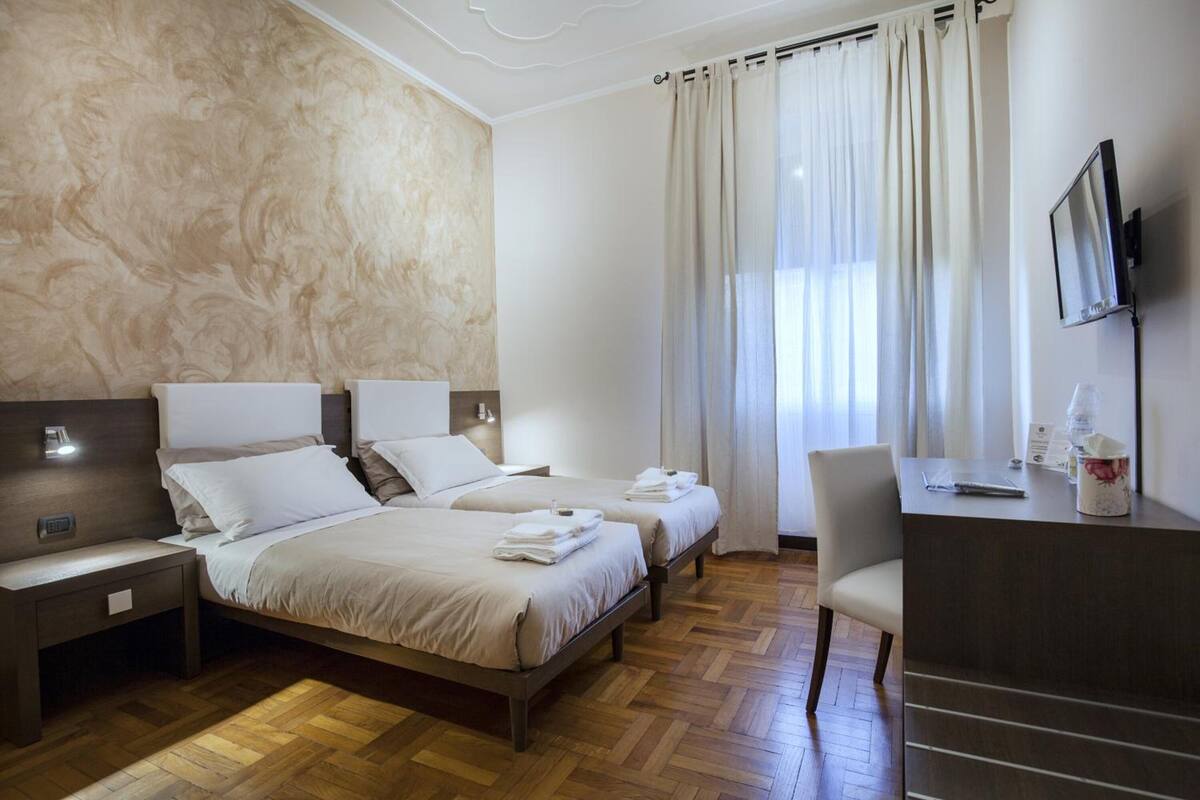A hotel room with two beds, a desk, a chair, a TV, and a large window with curtains.