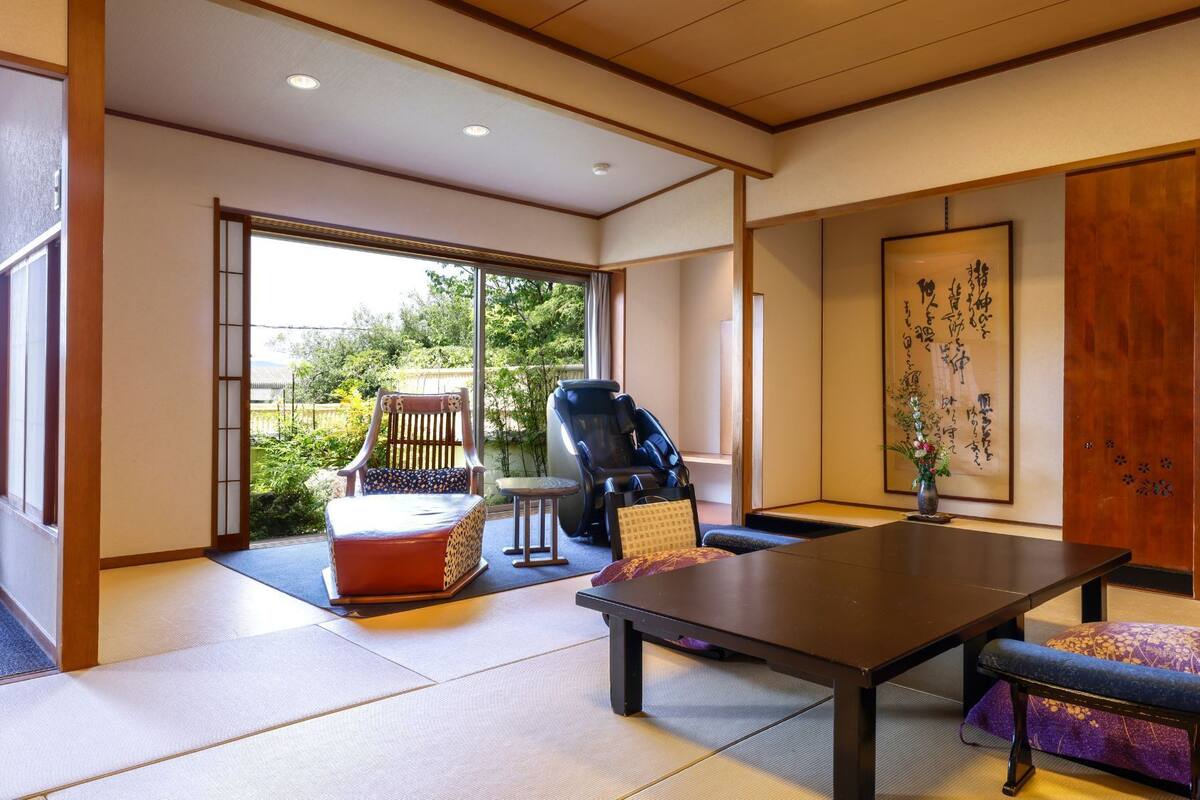 Una habitación japonesa tradicional con suelo de tatami, una mesa baja, una silla de masaje y un puertas correderas que dan a un jardín.