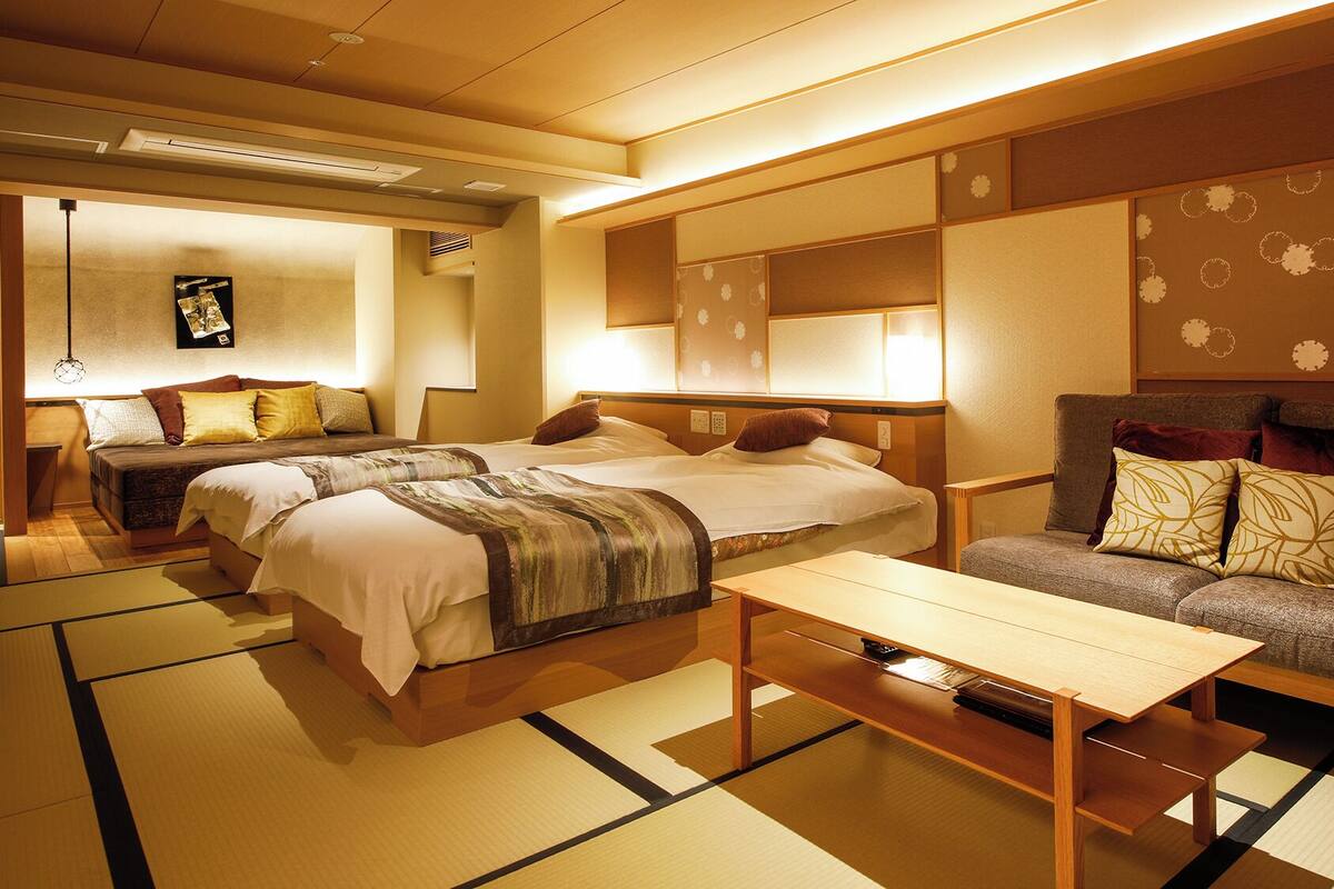 Habitación de hotel tradicional al estilo japonés, con suelo de tatami, un sofá, dos camas y una mesa.