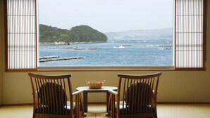 A room with tatami flooring, a low table with a woven basket, and two wooden chairs facing a large window overlooking a body of water and a distant island. - Sun Urashima Yuki no Sato (Toba)