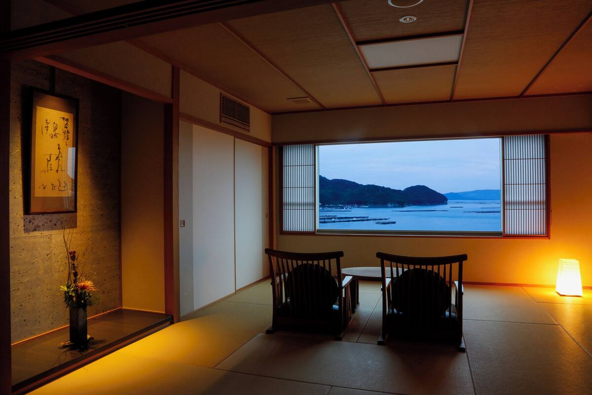 Um quarto japonês tradicional com tatames, uma mesa baixa, duas cadeiras, uma janela com persianas de bambu e um painel shoji fixado na parede.