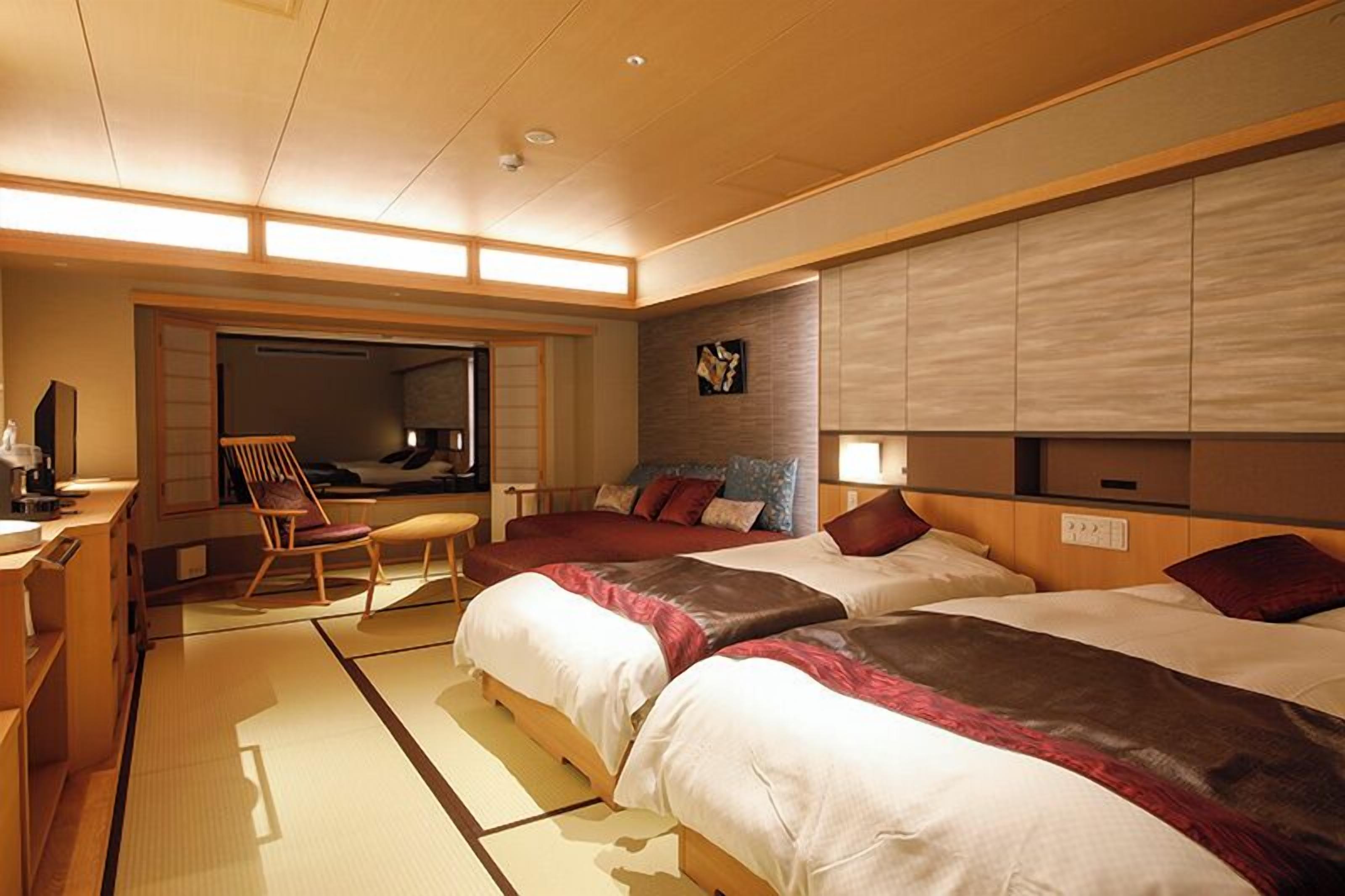 Ein Hotelzimmer mit zwei Betten, einer Sitzecke inklusive Sessel, einem kleinen Tisch und einem an der Wand befestigten Fernseher.