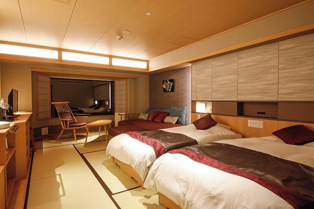 Quarto de hotel com duas camas, um espaço de estar com uma poltrona, uma mesinha e uma televisão fixada na parede.