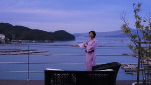 A person in a pink robe standing on a balcony overlooking a body of water with boats and distant hills. - Sun Urashima Yuki no Sato (Toba)