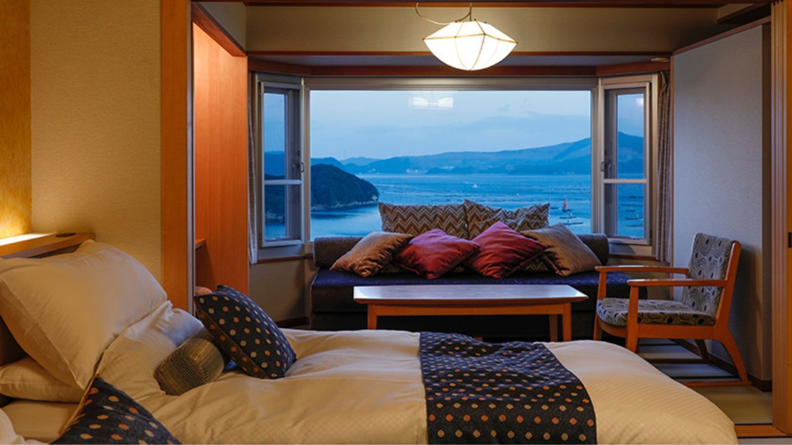 Ein Schlafzimmer mit Bett, einer Couch, einem Sessel, einem Tisch und Meerblick durch große Fenster.