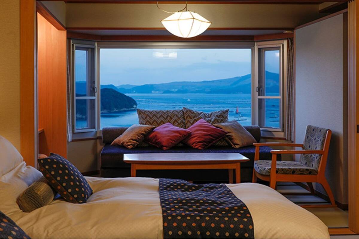 Un dormitorio con cama, sofá, sillón, mesa y vistas al mar a través de amplios ventanales.