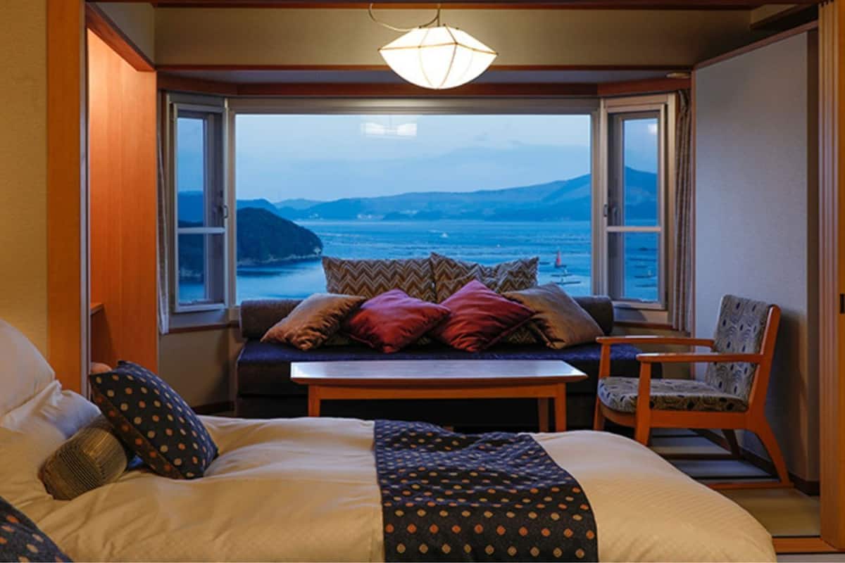 Une chambre à coucher comprenant un lit, un canapé, une chaise, une table et offrant une vue sur la mer grâce à de grandes fenêtres.